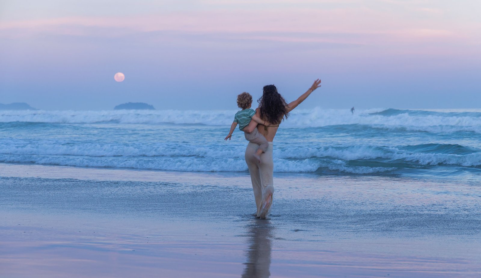Anita en la playa al atardecer, alzando a su hijo, con la luna sobre el mar